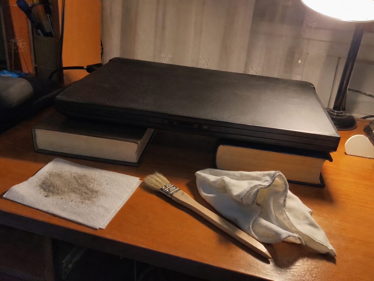Старый ноутбук приподнят на двух толстых книгах, рядом кисточка и салфетка с пылью