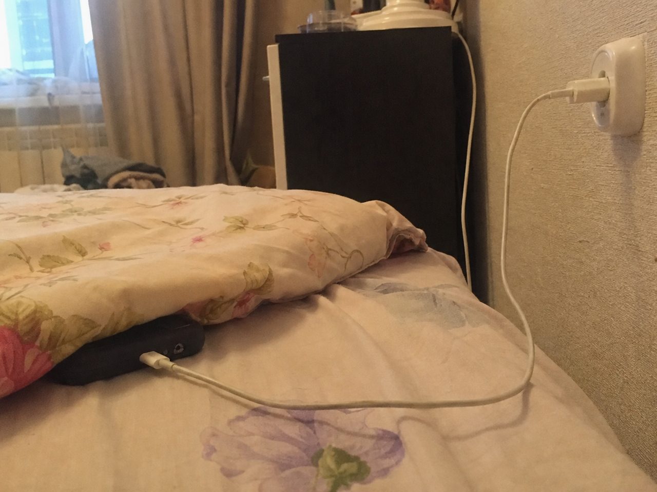 Телефон в плотном чехле на зарядке лежит под подушкой на неубранной кровати
