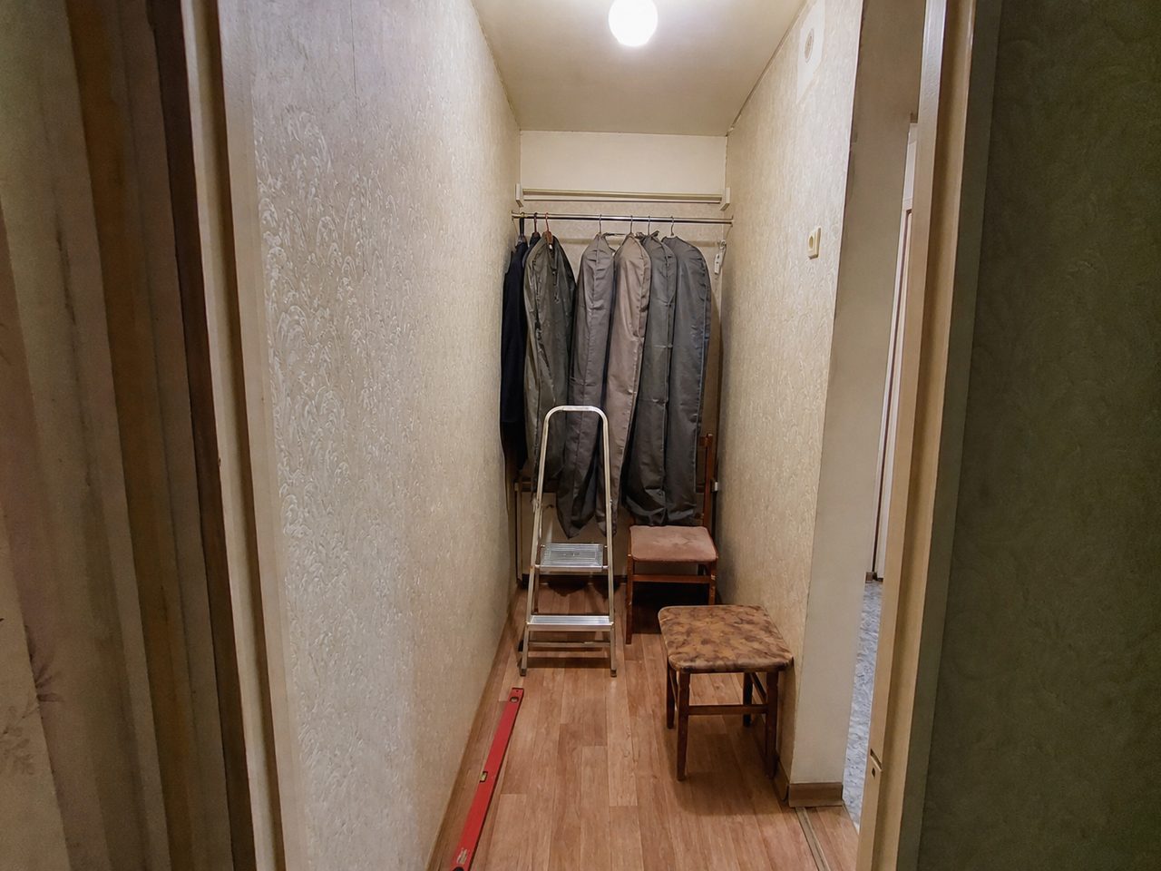 Узкая ниша в коридоре до ремонта: куртки в чехлах на штанге, стремянка и табуретки
