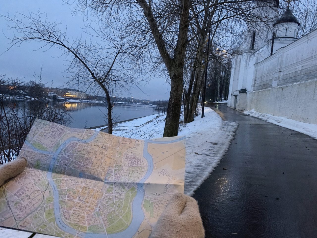 Заснеженный берег реки в старом городе, вдали белая церковная стена, руки в шерстяных перчатках держат бумажную карту