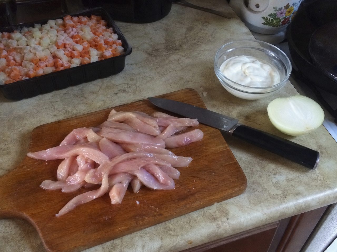 Разделочная доска с тонкими полосками куриного филе, нож, миска со сметаной и половинка лука